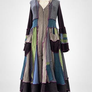May include: A long-sleeved, hooded coat featuring a patchwork design of knit fabric panels in shades of charcoal, slate gray, olive green, and teal. The garment includes a full-length front zipper, two side pockets, and exposed purple stitching.