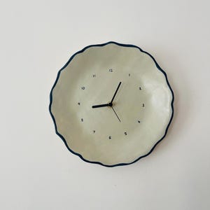 Puede incluir: Un reloj de pared redondo, blanquecino, con un borde ondulado azul oscuro. La esfera del reloj tiene números y manecillas negras. El diseño es minimalista y moderno, adecuado para la decoración del hogar. Diámetro aproximado de 25 cm.