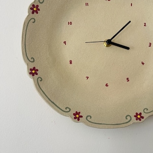 May include: A beige, scalloped-edge wall clock with a floral design. The clock face has red numerals and black hands. The clock is decorated with red flowers and green vines around the edge.