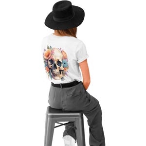 Puede incluir: Una camiseta blanca con un dise&ntilde;o de calavera floral en la espalda. La calavera est&aacute; rodeada de flores rosas, naranjas y azules. La persona que lleva la camiseta est&aacute; sentada en un taburete y lleva un sombrero negro.