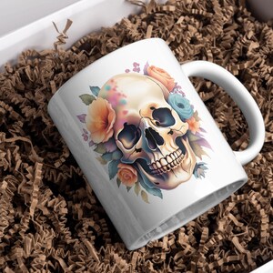 Puede incluir: Taza de cer&aacute;mica blanca con un dise&ntilde;o floral colorido que presenta una calavera con detalles pintados. La calavera est&aacute; rodeada de flores en tonos rosa, naranja y azul.