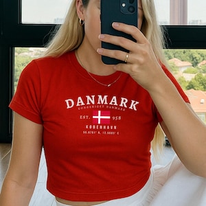 May include: A red cropped t-shirt with a white graphic that says "DANMARK" with the Danish flag and the text "EST. KOBENHAVN 1958". The model is wearing a white skirt.