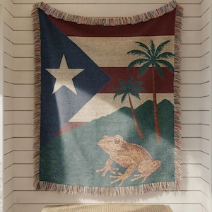 May include: A woven tapestry featuring the flag of Puerto Rico, with a white star on a blue triangle, and red and white stripes. The tapestry also depicts palm trees and a frog. The tapestry is hanging on a white wall.