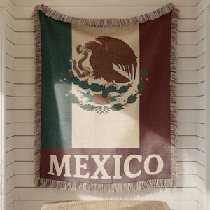 May include: A woven throw blanket featuring the Mexican flag design, with green, white, and red stripes. The center displays the Mexican coat of arms, with the word "MEXICO" in white block letters below. The blanket is fringed.
