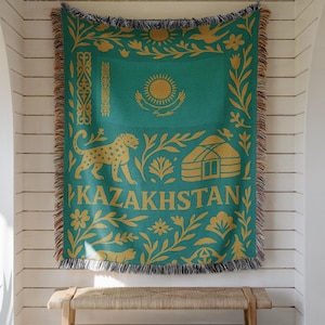 May include: A teal and gold woven throw blanket featuring the word "KAZAKHSTAN" and a design with a sun, leopard, and yurt. The blanket has a fringed edge and is hanging on a white wall.