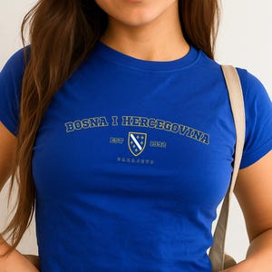 May include: A royal blue crop top with short sleeves. The shirt features the text "BOSNA I HERCEGOVINA" in a curved font, along with a crest and the year "1992". The shirt is paired with blue jeans.