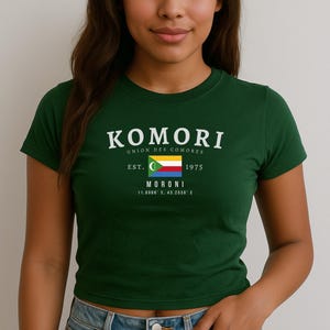 May include: A green t-shirt with a white graphic print that says "Komori Union des Comores Est. 1975 Moroni 11.6686° S, 43.2630° E" and features the flag of Comoros.