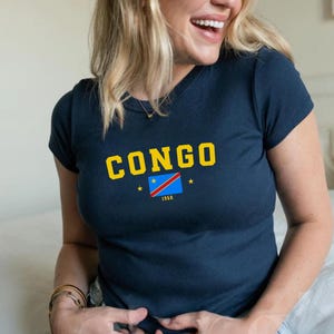 May include: A woman wearing a navy blue t-shirt with the text "CONGO" in yellow letters and a small flag of the Democratic Republic of the Congo. The shirt also has the year "1960" printed on it.