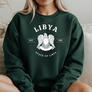 Può includere: Una felpa verde scuro con la scritta "LIBYA" in lettere bianche sopra un emblema bianco di un'aquila con le ali spiegate. Sotto l'emblema ci sono le parole "STATE OF LIBYA". La felpa ha maniche lunghe e girocollo.