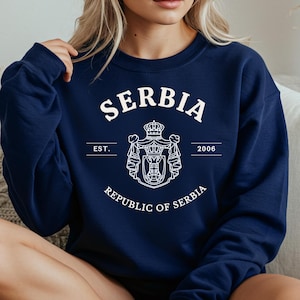 Puede incluir: Sudadera azul marino con la palabra "SERBIA" en blanco, arqueada sobre el pecho. Debajo, un emblema blanco con una corona y el texto "REPUBLIC OF SERBIA". También se ve el año "2006".