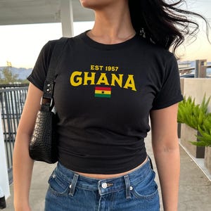 May include: A black t-shirt with the text "EST 1957 GHANA" in yellow letters and a small Ghanaian flag. The shirt is being worn by a person with dark hair and blue jeans.
