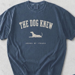 May include: A denim blue short-sleeve t-shirt featuring a graphic print in off-white. The text reads THE DOG KNEW and ARGOS OF ITHACA, accompanied by a line-art illustration of a dog lying down. The shirt has a crew neckline and a vintage wash.