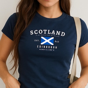 May include: Navy blue t-shirt with the word "SCOTLAND" in white, and a blue and white flag graphic. Below the flag is the word "EDINBURGH" and coordinates. The shirt has short sleeves and a crew neck.