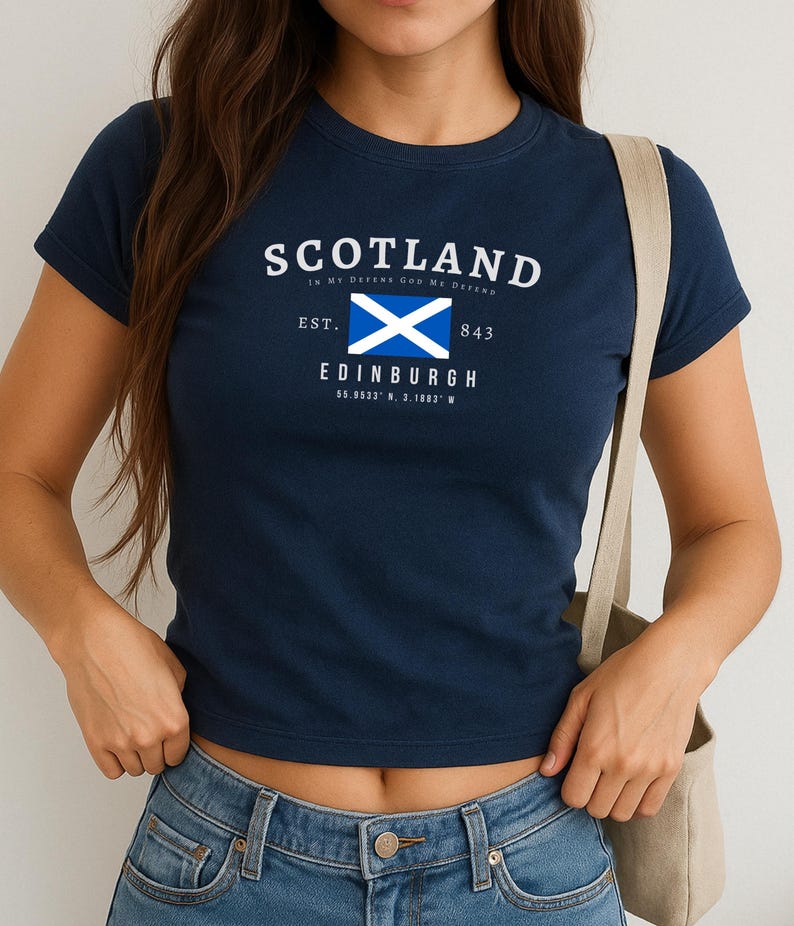 May include: Navy blue t-shirt with the word "SCOTLAND" in white, and a blue and white flag graphic. Below the flag is the word "EDINBURGH" and coordinates. The shirt has short sleeves and a crew neck.