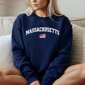 Könnte beinhalten: Ein marineblaues Sweatshirt mit dem Wort "MASSACHUSETTS" in weißer, bogenförmiger Schrift. Darunter befindet sich eine kleine amerikanische Flagge. Das Sweatshirt hat einen Rundhalsausschnitt.