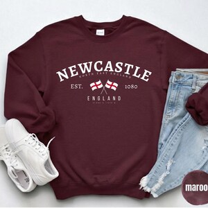 Puede incluir: Sudadera granate con la inscripci&oacute;n blanca "NEWCASTLE ENGLAND" y dos banderas cruzadas. La imagen incluye vaqueros de color azul claro y zapatillas blancas. La palabra "marr&oacute;n" se muestra en un c&iacute;rculo.