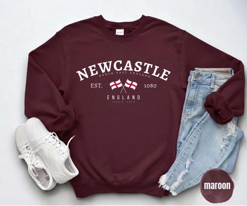 Puede incluir: Sudadera granate con la inscripci&oacute;n blanca "NEWCASTLE ENGLAND" y dos banderas cruzadas. La imagen incluye vaqueros de color azul claro y zapatillas blancas. La palabra "marr&oacute;n" se muestra en un c&iacute;rculo.