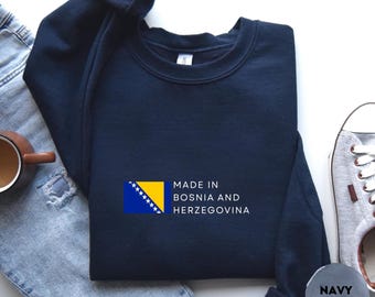 Könnte beinhalten: Marineblaues Sweatshirt mit dem Text "MADE IN BOSNIA AND HERZEGOVINA" und der Flagge von Bosnien und Herzegowina. Das Sweatshirt ist auf einer Jeans gefaltet. Eine marineblaue Farbmuster befindet sich in der unteren rechten Ecke.