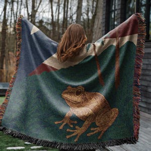 May include: A woven throw blanket featuring a frog design. The blanket has a dark blue corner with a white star and red stripe. The main body of the blanket is green with a brown frog illustration. The blanket has a fringed edge.
