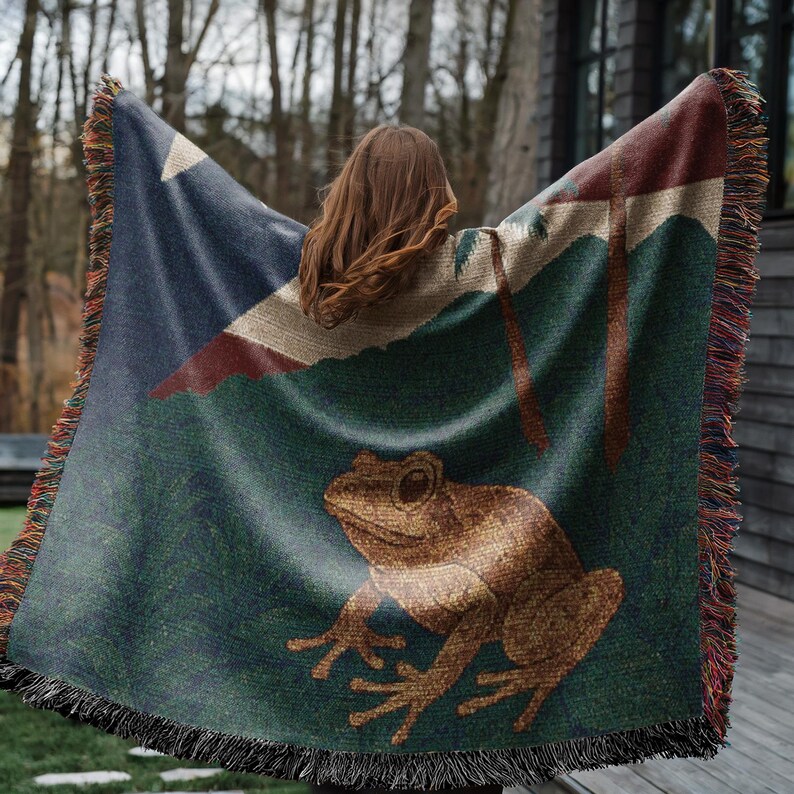May include: A woven throw blanket featuring a frog design. The blanket has a dark blue corner with a white star and red stripe. The main body of the blanket is green with a brown frog illustration. The blanket has a fringed edge.