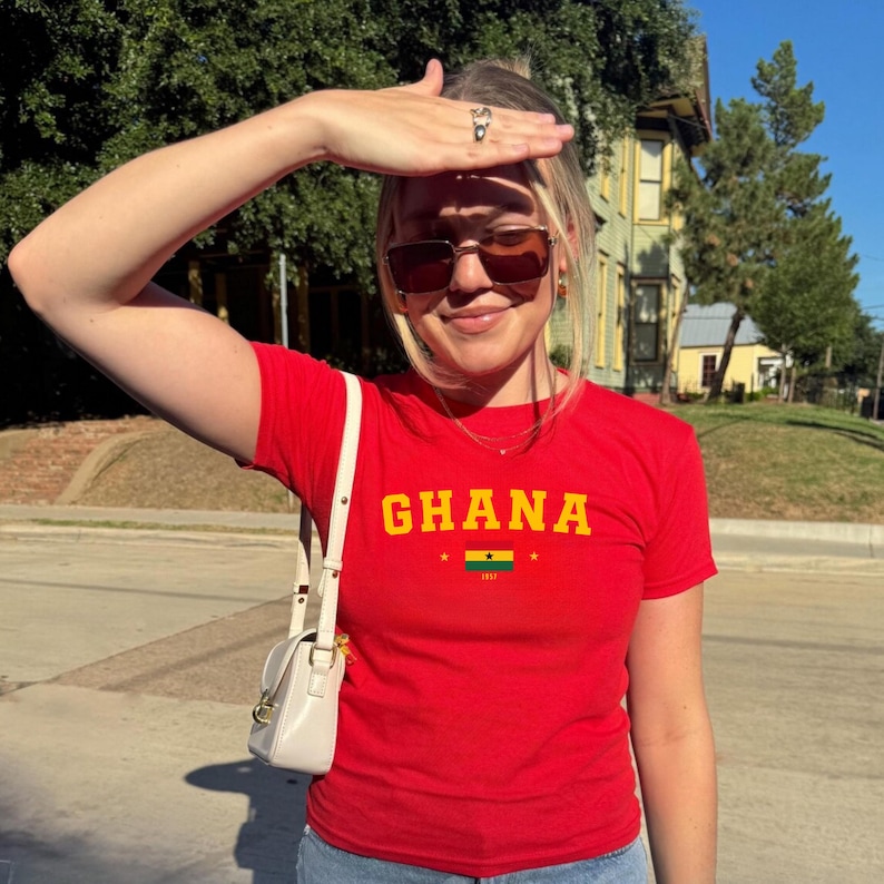 K&ouml;nnte beinhalten: Eine Frau tr&auml;gt ein rotes T-Shirt mit dem Wort "GHANA" in gelben Buchstaben und der ghanaischen Flagge, die auf das T-Shirt gedruckt ist. Sie tr&auml;gt eine Sonnenbrille und eine wei&szlig;e Umh&auml;ngetasche.