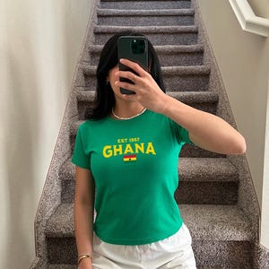 May include: A green t-shirt with short sleeves and a yellow and red graphic that says "EST. 1957 GHANA" with a small flag of Ghana below the text.