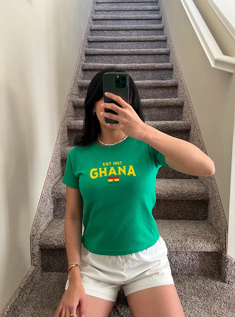 May include: A green t-shirt with short sleeves and a yellow and red graphic that says "EST. 1957 GHANA" with a small flag of Ghana below the text.