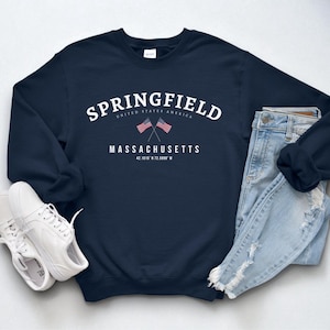 Puede incluir: Una sudadera azul marino con la palabra "SPRINGFIELD" en texto blanco arqueado, con "UNITED STATES AMERICA" debajo. Debajo, dice "MASSACHUSETTS" con dos banderas americanas cruzadas. Zapatillas blancas y vaqueros azul claro también están en la imagen.