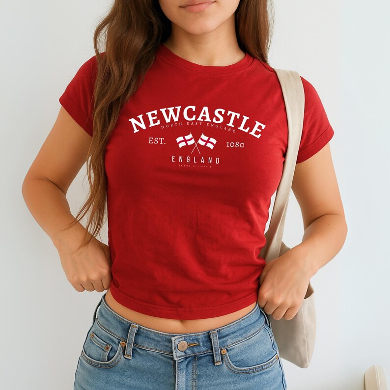 May include: A red t-shirt with white text that reads "Newcastle North East England" above two crossed flags and the word "England" below. The shirt is a crew neck style.