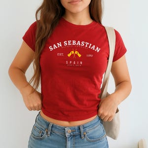 May include: A red t-shirt with the text "SAN SEBASTIAN" in white, arched across the top. Below, it reads "BASQUE COUNTRY EST. 1180 SPAIN" with two crossed flags. The shirt is short-sleeved and worn with blue jeans.