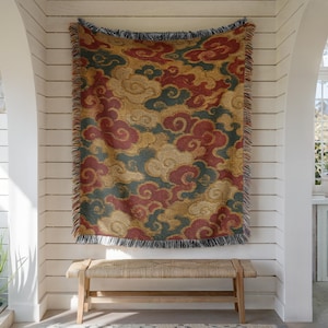 May include: A woven throw blanket featuring a repeating pattern of stylized clouds in shades of deep red, dark teal, and golden yellow. The rectangular textile has a fringed edge along the top and bottom, displaying a dense, textured weave.