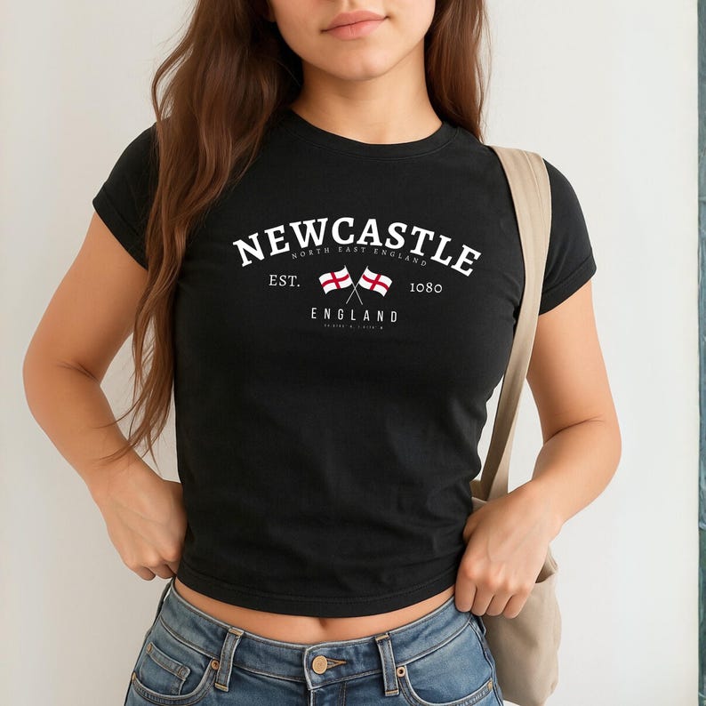 May include: Black t-shirt with white text that reads "Newcastle" above "North East England" and "England". Two crossed flags are in the center. The text "Est. 1080" is also present. The shirt is paired with blue jeans.
