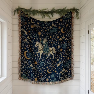 Könnte beinhalten: Eine gewebte Wohndecke mit dunkelblauem Hintergrund und einem wiederkehrenden Muster aus goldenen Sternen, Mondsicheln, Bergsilhouetten und wirbelnden Wolken. Eine zentrale Figur eines Zauberers auf einem weißen Pferd hält einen Sternenstab.