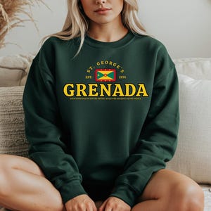 Może przedstawiać: Ciemnozielona bluza z napisem "GRENADA" w żółtych literach. Nad słowem znajduje się grafika flagi Grenady oraz napisy "ST. GEORGE'S EST. 1974". Bluza ma długie rękawy i okrągły dekolt.