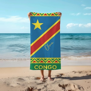 Puede incluir: Una toalla de playa azul con la bandera de la República Democrática del Congo. La toalla tiene una estrella amarilla, una franja diagonal roja y amarilla, y el nombre "Olivia" en cursiva. La palabra "CONGO" está impresa en la parte inferior.