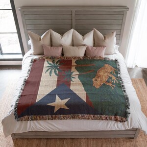 May include: A woven throw blanket featuring the flag of Puerto Rico, with a large star, blue triangle, and red stripes. The blanket also depicts a frog and palm trees in shades of green, brown, and beige. The blanket is fringed.