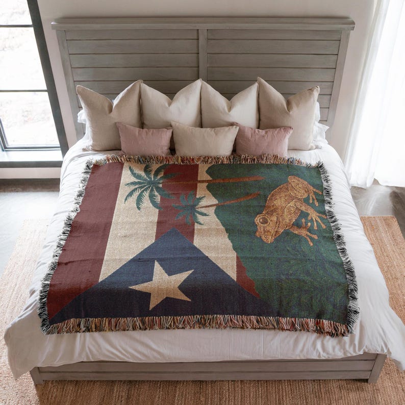 May include: A woven throw blanket featuring the flag of Puerto Rico, with a large star, blue triangle, and red stripes. The blanket also depicts a frog and palm trees in shades of green, brown, and beige. The blanket is fringed.