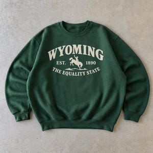 May include: A dark forest green crewneck sweatshirt featuring cream-colored text and a graphic. The design includes the word WYOMING, EST. 1890, and THE EQUALITY STATE, centered around a silhouette of a bucking horse and rider in the middle.