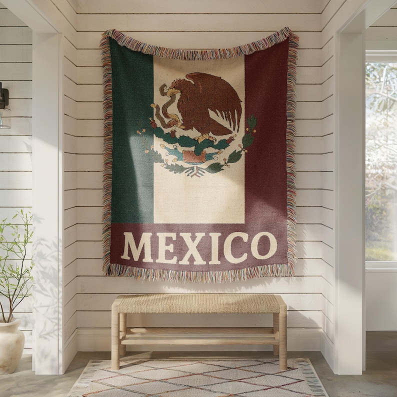 May include: A woven throw blanket featuring the Mexican flag design, with green, white, and red stripes. The center displays the Mexican coat of arms, with the word "MEXICO" in white block letters below. The blanket is fringed.