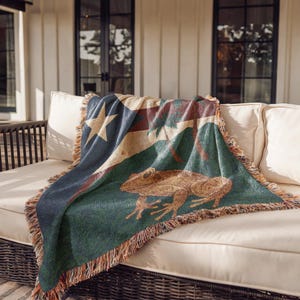 May include: A woven throw blanket featuring a large frog design in shades of brown, green, and blue. The blanket has a star and stripe pattern and is fringed along the edges. The blanket is draped over a white sofa.