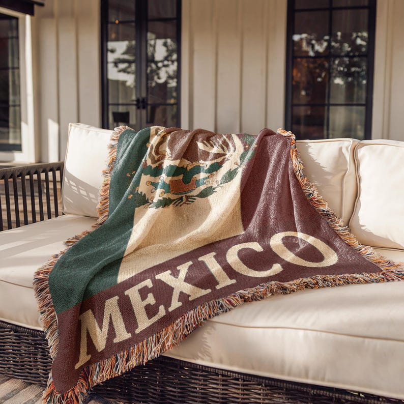 May include: Woven throw blanket featuring the Mexican flag design. The blanket is primarily green, white, and red, with the word "MEXICO" in large, off-white letters. The blanket has a fringed edge and is draped over a white couch.