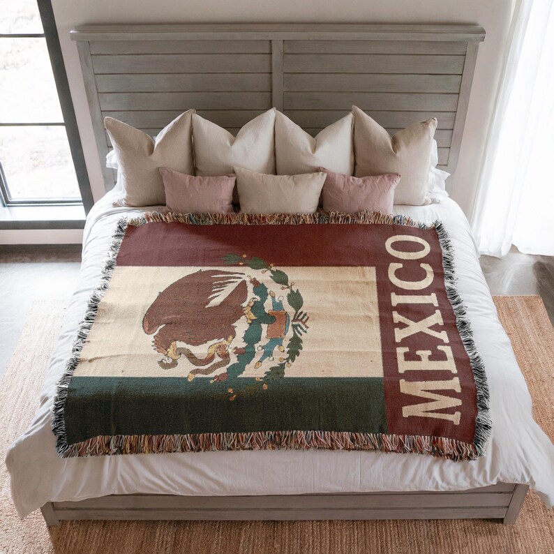 May include: A woven blanket featuring the Mexican flag design. The blanket displays the Mexican coat of arms, with a brown eagle and a snake, set against a cream, green, and burgundy background. The word "MEXICO" is vertically printed on the right side.