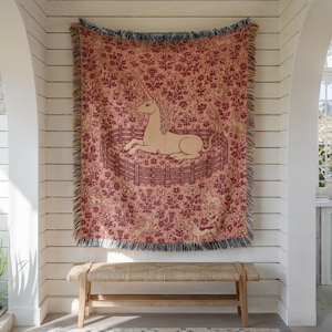 Puede incluir: Una manta tejida con un patrón repetitivo de un unicornio descansando dentro de una valla de madera, rodeado de conejos y motivos florales en tonos rosa empolvado, crema y rojo oscuro, con bordes deshilachados en todos sus lados.