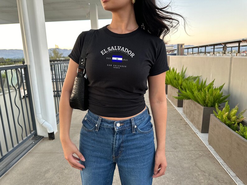 Może przedstawiać: Czarny T-shirt z białym nadrukiem flagi Salwadoru i tekstem "El Salvador" z napisami "San Salvador" i "Ameryka Środkowa" poniżej. Koszulkę nosi osoba w niebieskich dżinsach.