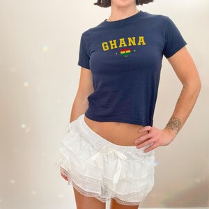 K&ouml;nnte beinhalten: Ein kurzes T-Shirt in Marineblau mit dem Wort "GHANA" in gelben Buchstaben und einer kleinen ghanaischen Flagge. Das Shirt wird mit einem wei&szlig;en, ger&uuml;schten Rock mit Spitzenbesatz kombiniert.