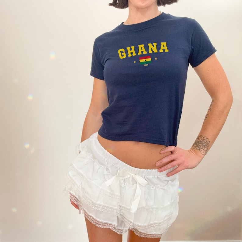 K&ouml;nnte beinhalten: Ein kurzes T-Shirt in Marineblau mit dem Wort "GHANA" in gelben Buchstaben und einer kleinen ghanaischen Flagge. Das Shirt wird mit einem wei&szlig;en, ger&uuml;schten Rock mit Spitzenbesatz kombiniert.