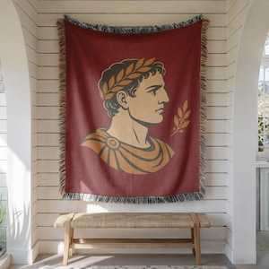 May include: A woven throw blanket in a deep maroon color featuring a central graphic of a Roman figure in profile wearing a laurel wreath. The design is rendered in shades of gold and dark gray, and the blanket has a decorative fringed edge.