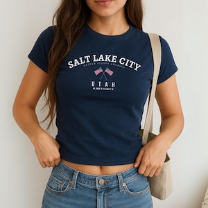 May include: A navy blue t-shirt featuring the white text "SALT LAKE CITY" arched across the top, with "UTAH" below. Two crossed American flags are in the center. The shirt also includes the coordinates 40.7600° N 111.8911° W.
