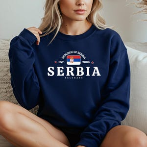 Pode incluir: Camisola azul marinho com a palavra "SERBIA" em branco, a bandeira sérvia e o texto "REPUBLIC OF SERBIA EST. 2006 BELGRADE". Gola redonda.