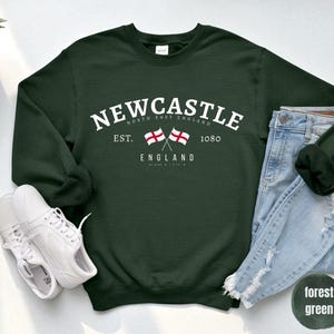 Puede incluir: Sudadera verde oscuro con la palabra "Newcastle" en blanco. Debajo, dos banderas cruzadas y la palabra "England". Zapatillas blancas y vaqueros azules junto a la sudadera.